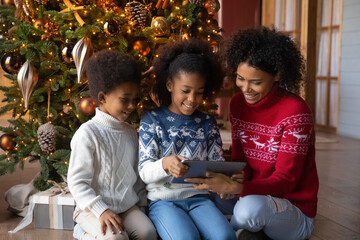 Happy young African American mother and two little kids sit near Christmas tree play on modern tablet. Smiling biracial mom and small children have fun using pad gadget on winter holidays at home.