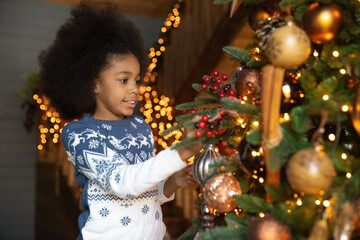Happy little African American girl have fun decorating beautiful Christmas tree at home alone. Smiling cute small biracial child kid prepare for New Year winter holidays celebration near fir-tree.
