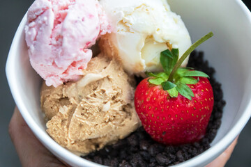 Hand holding a bowl of various ice cream flavor and crumbled chocolate cookies with strawberry on rustic background.