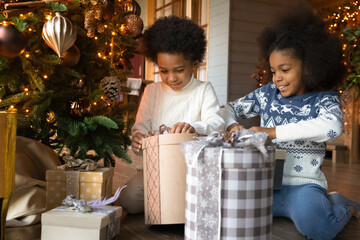 Excited two little African American children sit near fir-tree unpack wrapped Christmas presents...