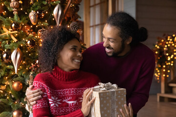 Fototapeta na wymiar Loving young African American husband congratulate excited wife with New Year winter holidays with gift. Smiling biracial man greet make surprise give present to beloved woman on Christmas.