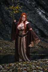 A full-length, plump red-haired girl in a historical medieval dress against the backdrop of gloomy...