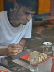 Fototapeta na wymiar View of a young guy eating fast food in the cafe seen through the window glass. 
