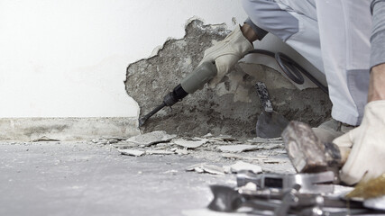 house renovation concept, construction worker breaks the old plaster of the wall with pneumatic air hammer chisel, close up with rubble and tools