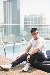 Fototapeta na wymiar Handsome young Asian man sitting on the rooftop near the swimming pool. Chinese guy, millennials life, lifestyle in the big city, Modern Asia