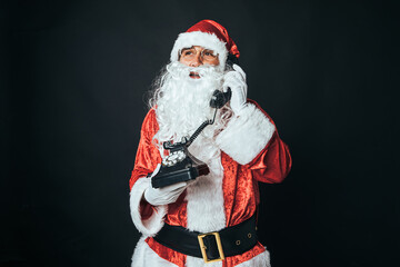 Man dressed as Santa Claus holding a retro telephone from the 60s, talking, on black background. Concept of Christmas, Santa Claus, gifts, celebration.