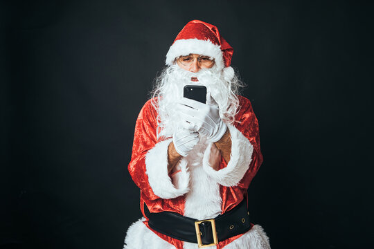 Man dressed as Santa Claus looking at his mobile phone, on black background. Concept of Christmas, Santa Claus, gifts, celebration.
