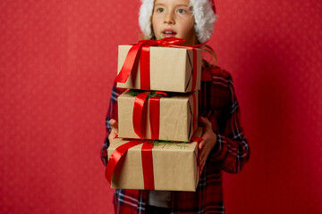 Funny little girl in santa hat with many Christmas gift boxes isolated on red background,
