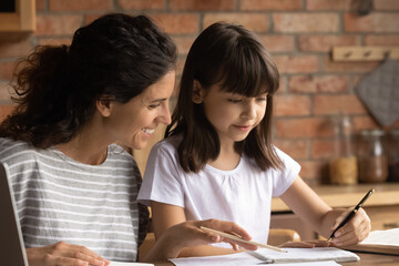 Happy mom helping primary schoolkid daughter to do homework. Nanny, sitter, teacher explaining school task to child, teaching kid to write in copybook at home. Homeschooling concept