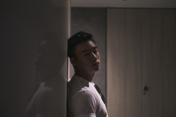 Portrait of handsome young pensive Asian man in white t-shirt. Standing near the wall and looking at camera with copy space