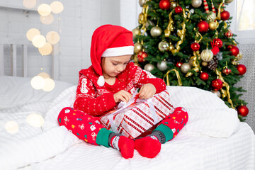 a child girl unpacks gifts at the Christmas tree in a red sweater and Santa Claus hat on New Year's...