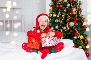 a child girl with gifts at the Christmas tree in a red sweater and Santa Claus hat on New Year's Eve or Christmas at home on a white bed screams with happiness