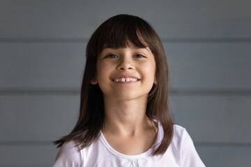 Happy cheerful black haired 7s girl looking at camera, smiling. Joyful school kid, child head shot portrait against grey wall, studio background. Childhood, childcare, family, youth concept.