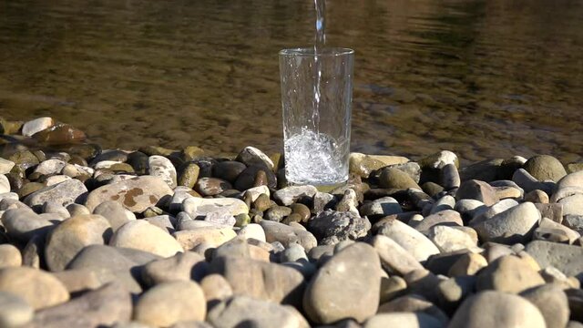 A high glassful standing on a pebble riverbank against a background of a flowing river is being filled with pure mineral water, slow motion.