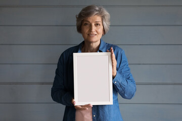 Smiling grey haired senior lady head shot. Positive 60s mature adult woman holding blank empty white picture frame, standing at studio background, looking at camera. Female portrait - Powered by Adobe