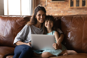 Happy little Latin daughter girl and young mom holding laptop, enjoying leisure time on sofa at home, looking at camera, smiling, distracting from watching movie, online shopping. Family portrait