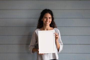 Happy pretty Hispanic 30s woman with curly hair holding blank empty white picture frame with copy space for text, standing at grey wall background, looking at camera. Head shot portrait - Powered by Adobe