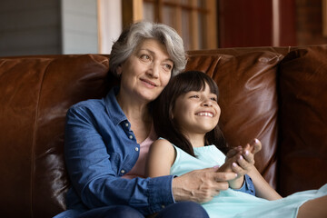 Happy grandmother hugging grand kid on couch at home, holding 7s girl in arms. Grey haired senior...