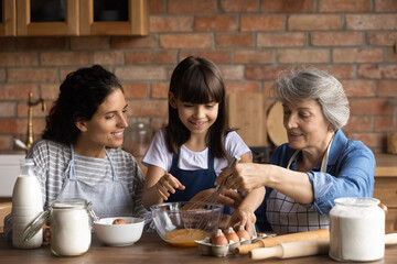 Happy Latin mom and grandma teaching girl to bake in kitchen. Mother, grandmother, kid cooking together, preparing dough for bakery food, mixing egg, flour in bawl on table. Family home activities