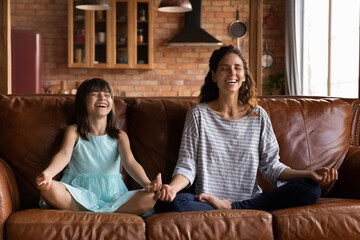 Happy young Hispanic mother and girl meditating on couch, sitting in lotus pose with zen hands,...