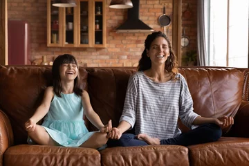 Fototapeten Happy young Hispanic mother and girl meditating on couch, sitting in lotus pose with zen hands, fingers, smiling with closed eyes. Mom and kid practicing yoga, mental exercise, mindfulness © fizkes