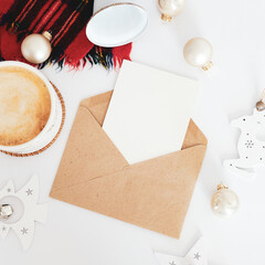 Craft paper envelope with Christmas letter mockup, cup of coffee, scarf, nordic Christmas decorations on white desk table. Scandinavian Christmas concept.