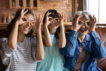 Happy granddaughter girl, mother, grandmother having fun at home together, enjoying leisure on sofa, looking at camera, smiling, laughing, making finger glasses. Family 3 generations portrait