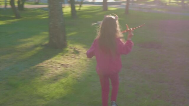 Happy long hair blonde little girl child in pink clothes with aeroplane running away from POV camera on outdoor sunlight day. Child play flying airplane, funny childhood.