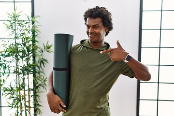 African man with curly hair holding yoga mat at studio looking confident with smile on face,...