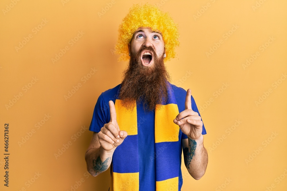 Sticker Redhead man with long beard football hooligan cheering game wearing funny wig amazed and surprised looking up and pointing with fingers and raised arms.