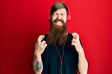 Redhead man with long beard listening to music using headphones gesturing finger crossed smiling with hope and eyes closed. luck and superstitious concept.