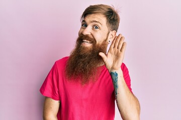 Redhead man with long beard wearing casual pink t shirt smiling with hand over ear listening and hearing to rumor or gossip. deafness concept.