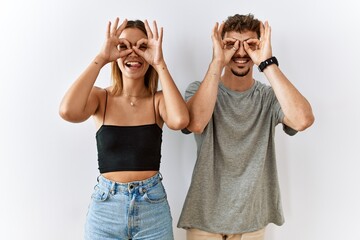 Young beautiful couple standing together over isolated background doing ok gesture like binoculars sticking tongue out, eyes looking through fingers. crazy expression.