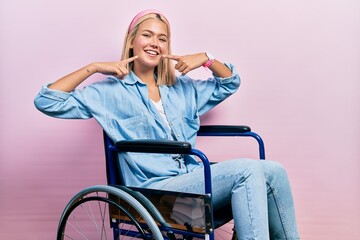 Beautiful blonde woman sitting on wheelchair smiling cheerful showing and pointing with fingers...