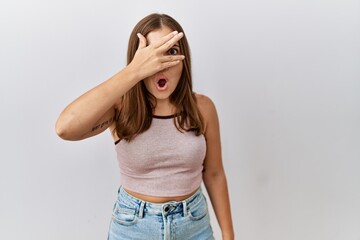 Young brunette woman standing over isolated background peeking in shock covering face and eyes with hand, looking through fingers with embarrassed expression.