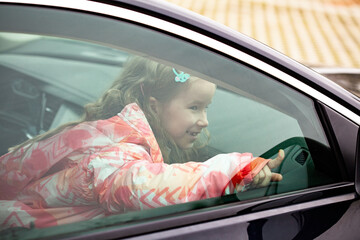 Little happy girl, child sitting inside the car seen from the outside. Young kid pointing outside the vehicle window, portrait, closeup. Means of transport, children traveling, transportation, road