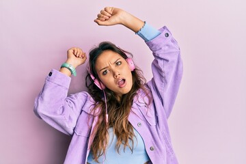 Young hispanic girl dancing and listening to music using headphones in shock face, looking skeptical and sarcastic, surprised with open mouth