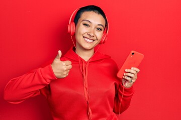 Beautiful hispanic woman with short hair using smartphone wearing headphones smiling happy and positive, thumb up doing excellent and approval sign