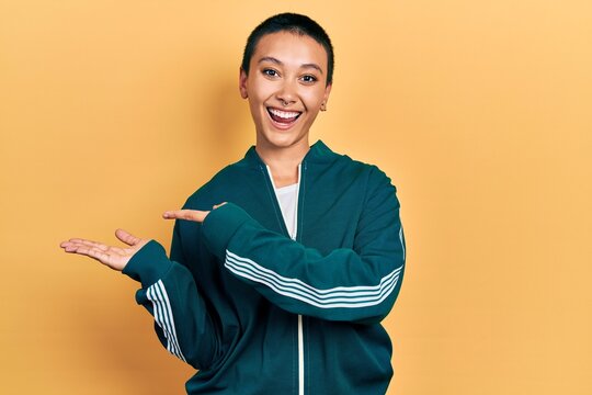 Beautiful hispanic woman with short hair wearing sporty jacket amazed and smiling to the camera while presenting with hand and pointing with finger.