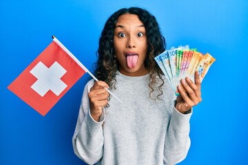 Young latin woman holding switzerland flag and swiss franc banknotes sticking tongue out happy with...