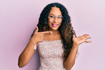 Young latin woman wearing casual clothes and glasses amazed and smiling to the camera while presenting with hand and pointing with finger.