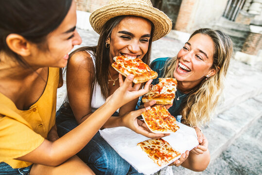 Three young female friends sitting outdoor and eating pizza - Happy women having fun enjoying a day out on city street - Happy lifestyle and tourism concept