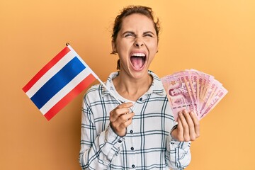 Young brunette woman holding thailand flag and baht banknotes angry and mad screaming frustrated...
