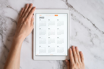 tablet computer with an open app of calendar for 2022 year in a womans hands on a gray marble...
