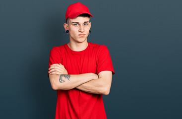 Young caucasian boy with ears dilation wearing delivery uniform and cap skeptic and nervous, disapproving expression on face with crossed arms. negative person.