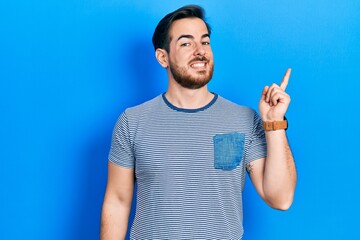 Handsome caucasian man with beard wearing casual striped t shirt with a big smile on face, pointing with hand and finger to the side looking at the camera.