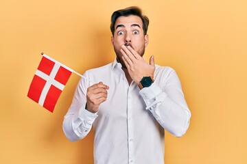 Handsome caucasian man with beard holding denmark flag covering mouth with hand, shocked and afraid...