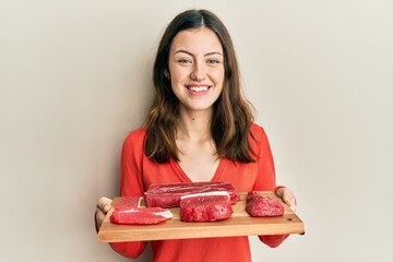 Young brunette woman holding board with raw meat smiling with a happy and cool smile on face....