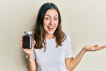 Young brunette woman holding bottle with coffee beans celebrating achievement with happy smile and winner expression with raised hand
