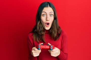 Young brunette woman playing video game holding controller afraid and shocked with surprise and amazed expression, fear and excited face. © Krakenimages.com